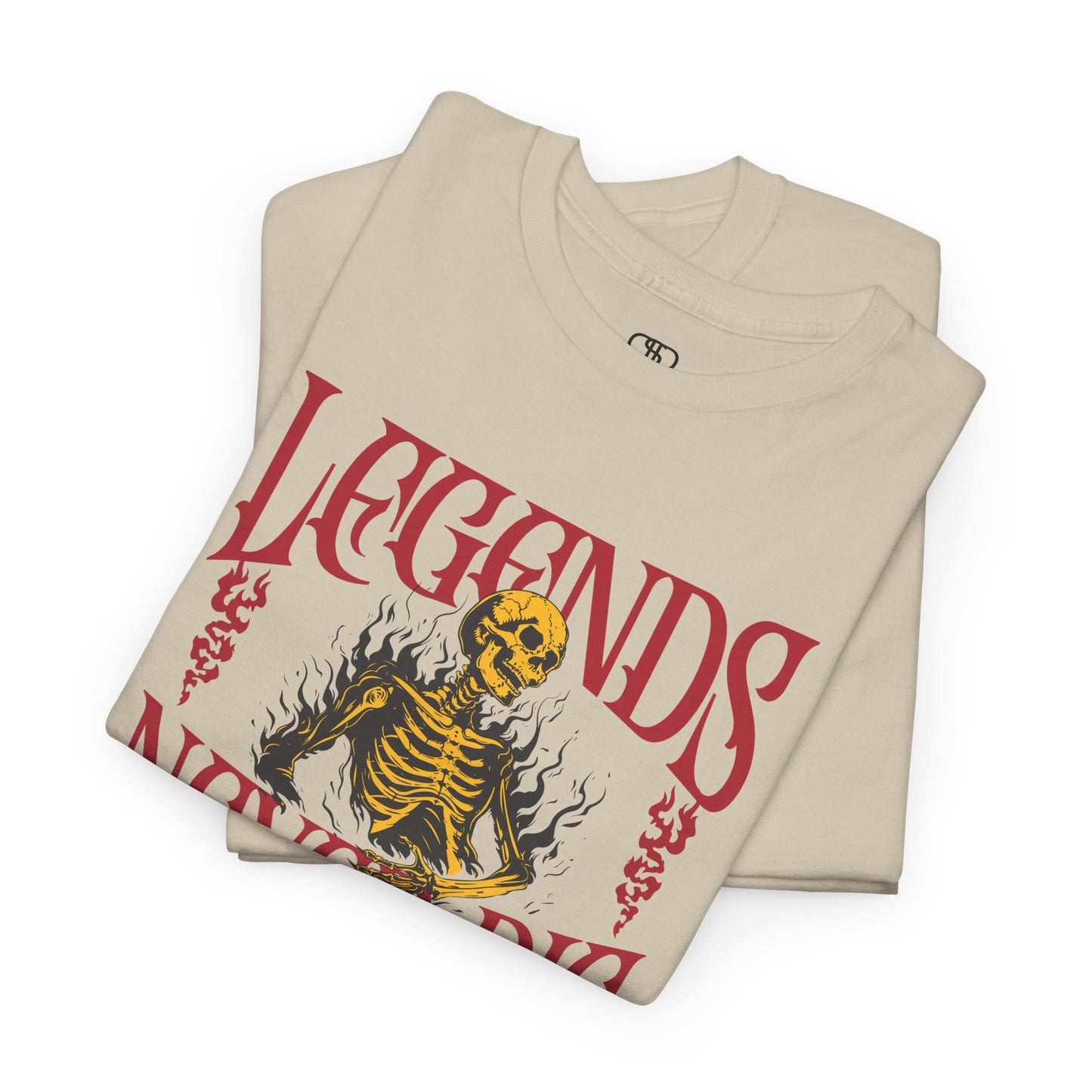 A folded sand t-shirt with a graphic of a defiant skeleton engulfed in flames. The words “Legends Never Die” are prominently displayed, with a smaller quote and the word “Immortal” included in the design.