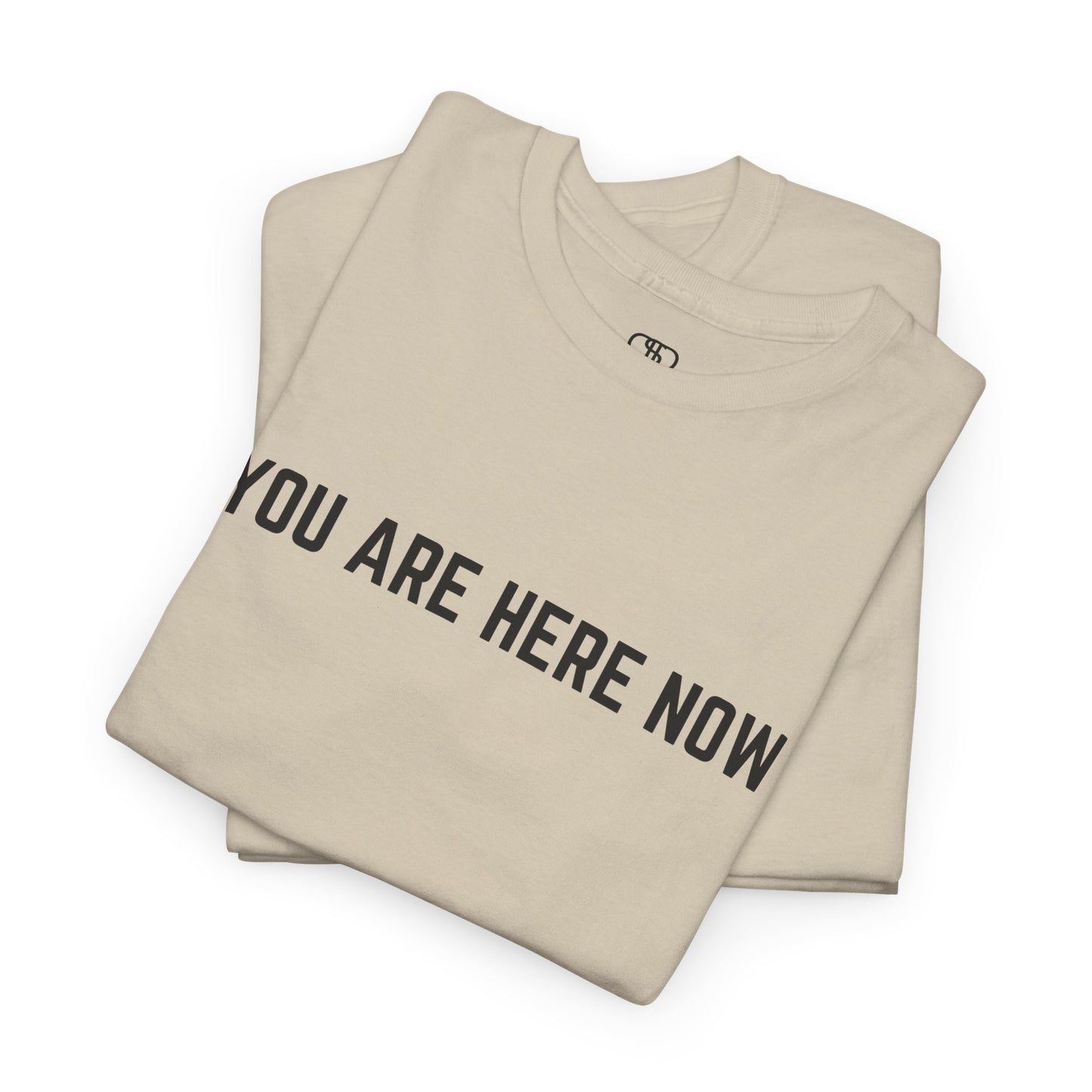 A folded sand t-shirt with the words "You are Here Now."