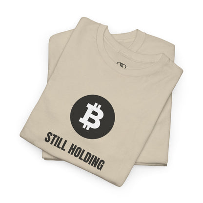 A folded sand T-shirt with a Bitcoin emblem and "Still Holding" text in black.