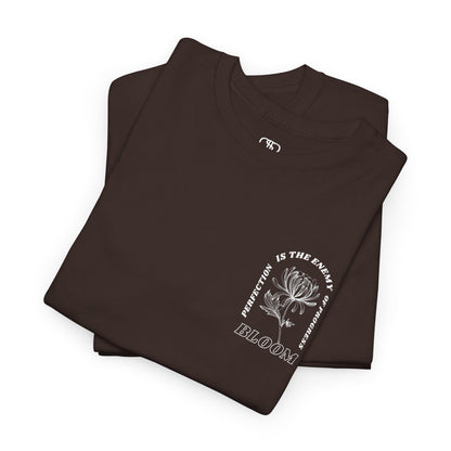 A [folded dark brown] t-shirt with a central flower design and the words “Perfection is the enemy of progress” and “Bloom” underneath it the phrase "Consistent effort breeds greatness," featured on the back of the t-shirt.