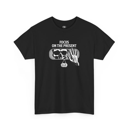 A t-shirt graphic of a car's rearview mirror reflecting a skeleton's face. A living hand reaches toward the mirror, which has dice dangling from it. The phrase “Focus on the present” is part of the design.