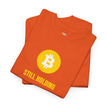 A folded orange  T-shirt with a Bitcoin emblem and "Still Holding" text in gold.