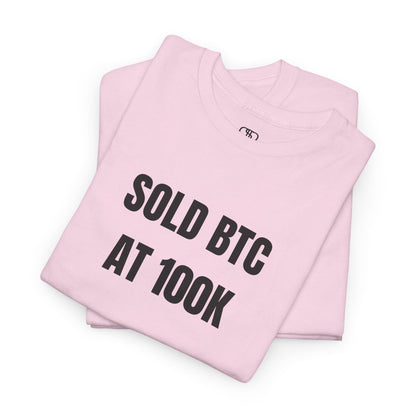 A folded light pink T-shirt with bold text "SOLD BTC At 100K" on the front.