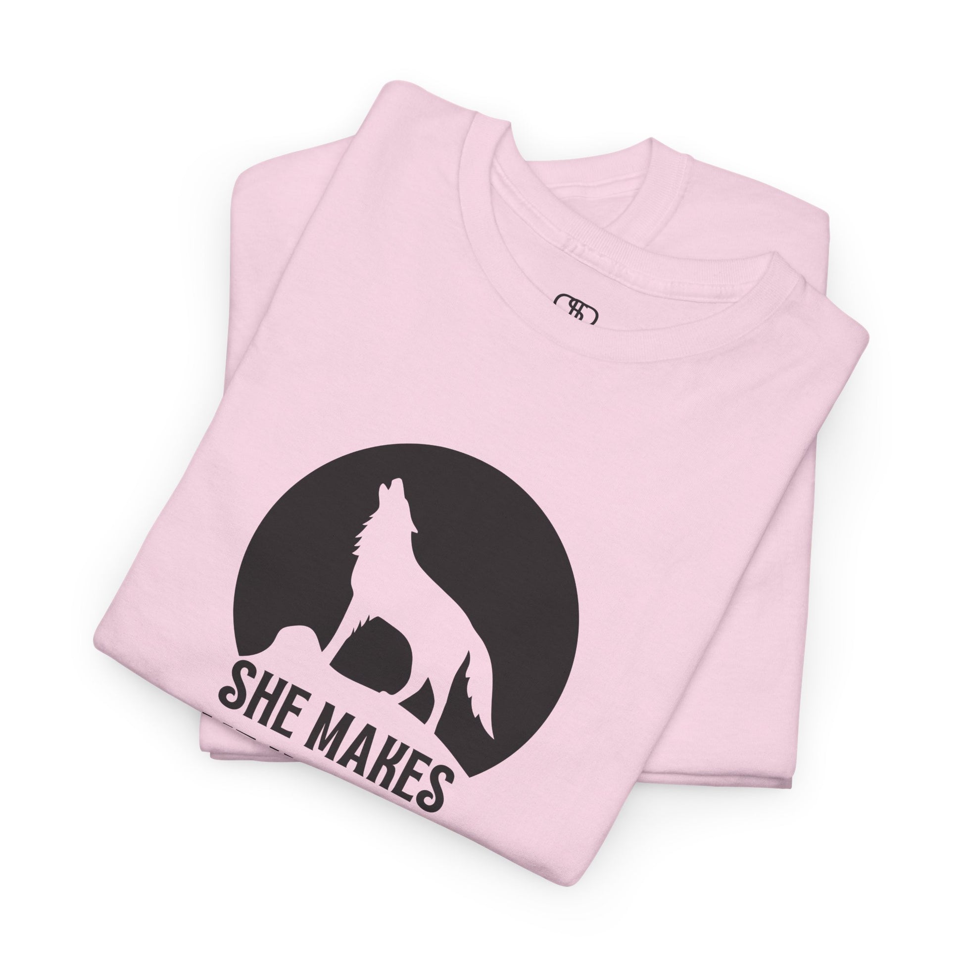 A folded light pink T-shirt with a wolf silhouette howling at the moon on a cliff, and "She makes me HOOWWL" text.