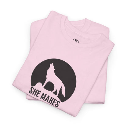 A folded light pink T-shirt with a wolf silhouette howling at the moon on a cliff, and "She makes me HOOWWL" text.
