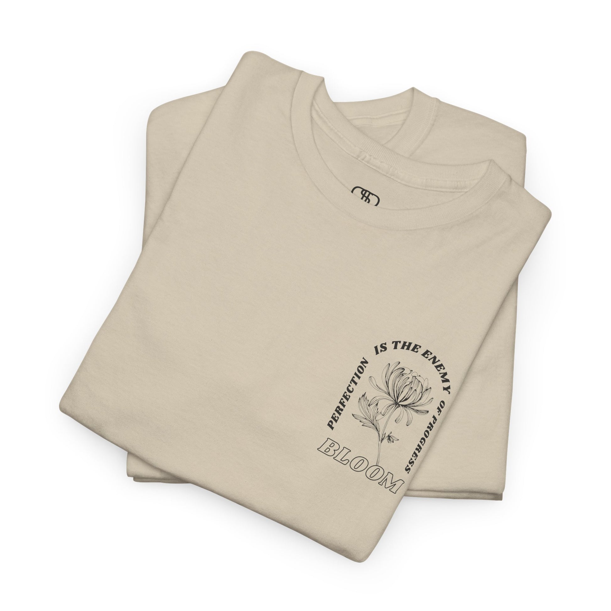 A [folded sand] t-shirt with a central flower design and the words “Perfection is the enemy of progress” and “Bloom” underneath it the phrase "Consistent effort breeds greatness," featured on the back of the t-shirt.