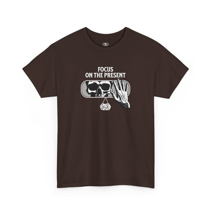 A t-shirt graphic of a car's rearview mirror reflecting a skeleton's face. A living hand reaches toward the mirror, which has dice dangling from it. The phrase “Focus on the present” is part of the design.