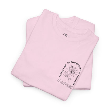 A [folded light pink] t-shirt with a central flower design and the words “Perfection is the enemy of progress” and “Bloom” underneath it the phrase "Consistent effort breeds greatness," featured on the back of the t-shirt.