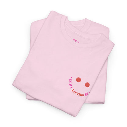 A folded pink t-shirt with a retro smiley face on the front and the phrase “In my lifting Era” in a creative retro design on the back.