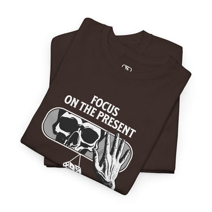 A t-shirt graphic folded of a car's rearview mirror reflecting a skeleton's face. A living hand reaches toward the mirror, which has dice dangling from it. The phrase “Focus on the present” is part of the design.