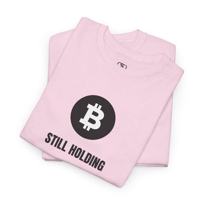 A folded light pink T-shirt with a Bitcoin emblem and "Still Holding" text in black.