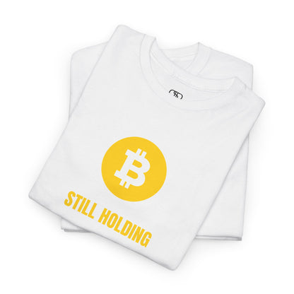 A folded white  T-shirt with a Bitcoin emblem and "Still Holding" text in gold.
